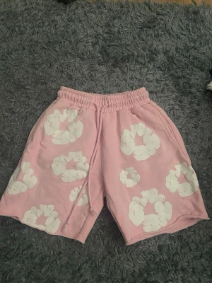 Rosa shorts med vita blommor - Säljer ett par rosa shorts med vita blommönster. Shortsen har resår i midjan och snörning framtill för extra komfort. De är tillverkade i mjukt material och har en bakficka. Perfekta för sommarens alla äventyr!
