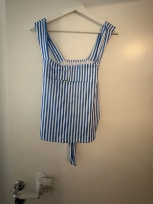 Blåvit randig linnetopp med knyt - Säljer en blåvit randig linnetopp med breda axelband och knyt i ryggen. Toppen har raka linjer och är ärmlös, perfekt för sommardagar. Snygg att styla med jeans eller kjol för en fräsch look. Använd 1 gång men är så så fin 
