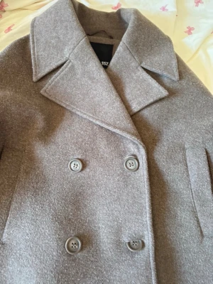 Ljusbrun dubbelknäppt ullkappa - Stilren ljusbrun kappa i ullmix med bred krage och dubbelknäppning framtill. Klassisk modell med raka fickor och stora knappar, perfekt för kyliga dagar. Snygg och tidlös design som passar till det mesta.