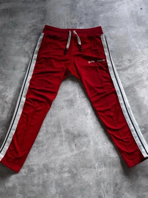 Röda mjukisbyxor Palm Angels - Snygga röda mjukisbyxor från Palm Angels med vita sidoränder och dragsko i midjan. Byxorna har en sportig look med smal passform och vita detaljer. Perfekta för en avslappnad streetwear-stil. Det finns några små fläckar på byxorna men dem går säkert bort i tvätten👍priset är ej hugget i sten så var inte rädda att komma med förslag🤝 Det står Strl L i byxorna men dem passar nog bättre till S och M