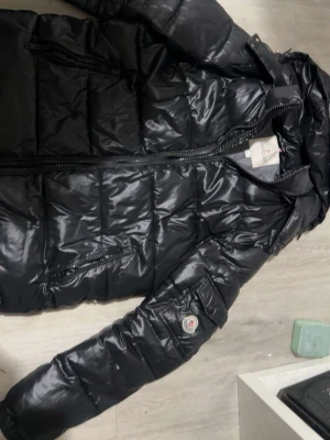 Svart dunjacka från Moncler - Snygg svart dunjacka från Moncler med glansig finish och klassisk quiltad design. Jackan har dragkedja framtill, fickor med dragkedja och Moncler-logga på ärmen. Perfekt för kalla vinterdagar och riktigt varm Dragkedjan på jackan fungerar inte.