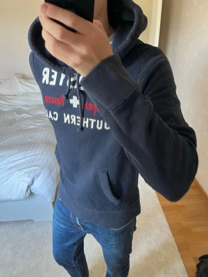 Mörkblå Hollister hoodie med tryck - Säljer en sjukt fet vintage hoodie från Hollister. Hoodien är i ett väldigt bra skick utan defekter och storleken är M! ✌🏼😁——————————————————————————                                                    📐| Storlek: M                                                             👖| Märke: Hollister                                                🔎| Skick: Mycket bra!                                                     ✍🏻| Skriv om du har några frågor