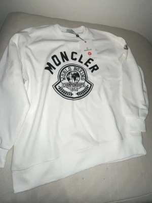 Vit Moncler sweatshirt med tryck - Snygg vit sweatshirt från Moncler med svart tryck framtill där det står 'World Boxing Championships 1952'. Tröjan har rund halsringning, långa ärmar och ribbade muddar. Perfekt för en sportig och trendig look.