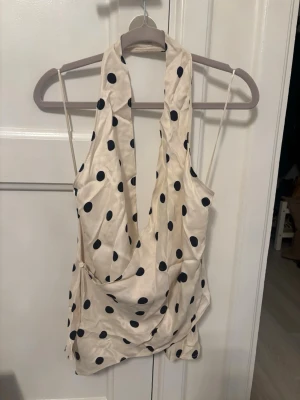 Prickig omlott halterneck topp - Snygg vit omlottopp med svarta prickar och halterneck. Toppen har djup urringning och tunna axelband, vilket ger en trendig och luftig look. Materialet ser ut att vara glansigt och liknar satin. Perfekt för dig som vill sticka ut med ett coolt mönster.