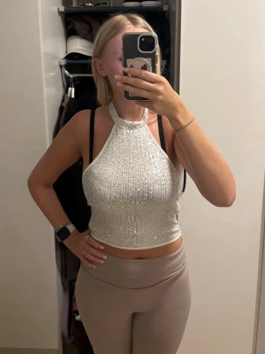 Glittrig silver halterneck topp - Säljer en glittrig silverfärgad halterneck topp med croppad passform. Toppen är ärmlös och har ett snyggt paljettmönster som fångar ljuset. Perfekt för dig som vill sticka ut och glänsa lite extra. Den är även öppen i ryggen 
