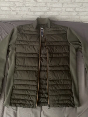 Olivgrön  cardiganjacka - Snygg olivgrön cardiganjacka med quiltad front och rygg samt mjuka fleeceärmar. Jackan har hög krage och dragkedja framtill. Perfekt för lager-på-lager och ger en sportig look. Materialmix av vadderad polyester och fleece.