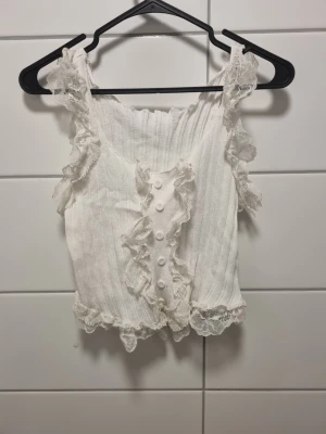 Vit spetsdetaljerad linnetopp - Supersöt vit linnetopp med spetsdetaljer längs axelbanden och nederkanten. Toppen har dekorativa knappar framtill, volangdetaljer och är ribbad för en extra snygg look. Perfekt för dig som gillar romantisk och feminin stil.