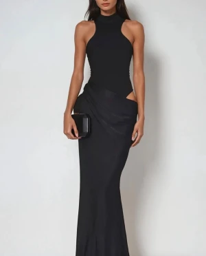 Svart långklänning med cut-out - Stilren svart långklänning med hög hals och ärmlös design. Klänningen har en tight passform upptill och en elegant cut-out detalj vid midjan. Materialet ser ut att vara mjukt och följsamt, perfekt för en trendig look.