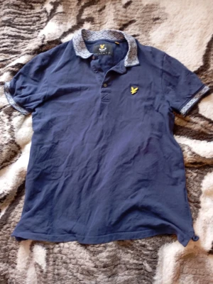 Marinblå piké med mönstrad krage - Snygg marinblå pikétröja från Lyle & Scott med korta ärmar och unik mönstrad krage samt ärmslut. Tröjan har knappslå framtill och den klassiska gula loggan på bröstet. Tillverkad i mjuk bomull för skön känsla. Inte mycket använd, men tyvärr har mittenknappen lossnat. Men det finns en knapp med om man skulle vilja sy fast en. Jag har bara inte orkat det. Pris kan diskuteras🙌. Mvh Rasmus.