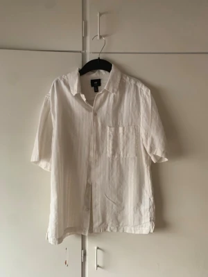 Vit kortärmad linneskjorta - Säljer en vit skjorta i linne med korta ärmar och klassisk krage. Skjortan har en bröstficka och knäppning framtill. Perfekt för varma dagar och enkel att styla till olika outfits.