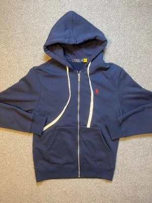 Marinblå hoodie från Polo Ralph Lauren - Snygg marinblå hoodie med dragkedja från Polo Ralph Lauren. Har vit dragsko i huvan, två fickor framtill och den klassiska röda loggan broderad på bröstet. Tillverkad i mjuk bomullsblandning, perfekt för chill dagar eller när du vill vara casual.