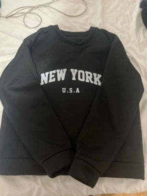 Svart sweatshirt med New York-tryck - Cool svart sweatshirt med vit text 'NEW YORK U.S.A' på bröstet. Klassisk rund halsringning och ribbade muddar vid ärmar och nederkant. Materialet känns mjukt och bekvämt.