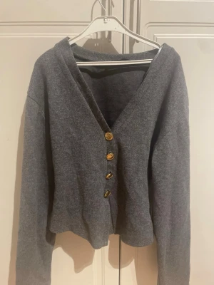 Grå kofta från Zara med guldknappar - Mysig grå kofta från Zara med fyra snygga guldknappar framtill. Koftan har v-ringning, långa ärmar och är tillverkad i ett mjukt stickat material. Perfekt att slänga över en topp för en chill look. 