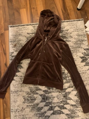 Brun velourhoodie med dragkedja - Mjuk och skön brun hoodie i velour med dragkedja framtill och två fickor. Justerbar huva med snören och ribbade muddar vid ärmar och nederkant. Perfekt för en avslappnad och trendig look.