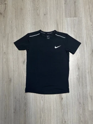Svart Nike Miller T-Shirt - Säljer en svart kortärmad löpartröja från Nike med vita detaljer på axlarna och klassisk Nike Swoosh på bröstet. Tillverkad i ett lätt och snabbtorkande material som passar perfekt för träning och löpning.