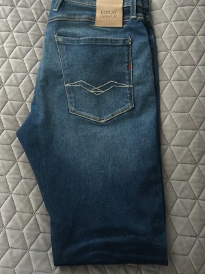 Blå Replay Hyperflex jeans - Snygga blå jeans från Replay Hyperflex med klassisk femficksmodell och vita kontrastsömmar. Modell anbass.Jeansen har en smal passform och stretchigt material som ger extra komfort. Bakfickan har unik broderad detalj och Replay-lapp i midjan.