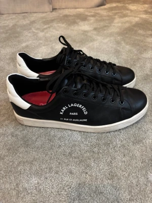 Svarta Karl Lagerfeld sneakers - Snygga svarta sneakers från Karl Lagerfeld med vita detaljer och vit sula. Skorna har svart snörning, låg profil och texten 'KARL LAGERFELD PARIS 21 RUE ST-GUILLAUME' på sidan. Tillverkade i slätt läder för en clean och modern look. Skorna är använda 1 gång.