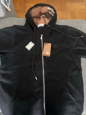 Svart hoodie med dragkedja och huva - Snygg svart hoodie med dragkedja, huva med dragsko och klassisk ficka framtill. Insidan av huvan har ett rutigt mönster och bröstet pryds av en diskret broderad logga. Perfekt för en chill och stilren look, finns i S M L 
