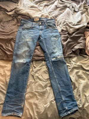 Blå slitna raka jeans - Säljer ett par blå jeans med raka ben och slitna detaljer på både fram- och baksida. Jeansen har klassisk femficksdesign och ljus tvätt med snygga slitningar på knän och fickor. Perfekta för en avslappnad och trendig look. Slim fit W31L32