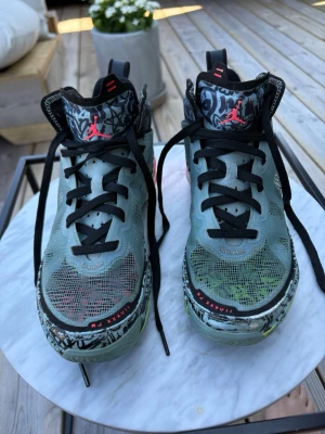 Jordan sneakers/basketskor med grafiskt mönster strl 39 - Unika Jordan sneakers med högt skaft, svart och grått grafiskt mönster, svarta skosnören och detaljer i rött och grönt. Ovandelen är i mesh och syntetmaterial, med färgglada innersulor och tydlig Jumpman-logga på plösen. Perfekta för dig som vill sticka ut. Bra skick, sparsamt använda. Lite slitage på framsidan av skon. Se bild! Ny pris är 2200 kr men jag säljer de för 1200. De är använda högst 15 gånger då de var på gränsen för att vara för små när jag köpte dem.