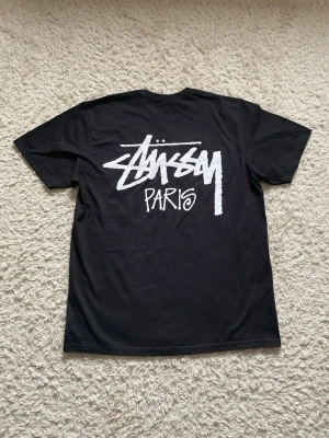 Stüssy Paris T-shirt  - Svart t-shirt från Stüssy med stort vitt Stüssy Paris-tryck på ryggen och mindre logga på bröstet. Klassisk passform och rund hals. Perfekt för dig som gillar streetwear och vill ha en clean look.