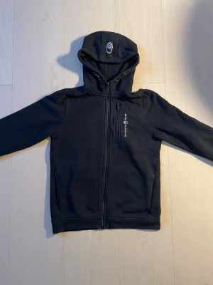 Svart hoodie med dragkedja och huva - Snygg svart hoodie med dragkedja framtill och huva. Har en bröstficka med dragkedja och tryckt logga på vänster sida. Tillverkad i ett mjukt och stretchigt material som passar perfekt till chill eller streetwear.
