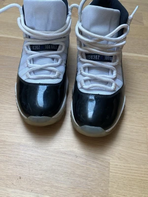 Nike Air Jordan 11 svartvita sneakers - Säljer ett par Nike Air Jordan 11 sneakers i svart och vitt. Skorna har en blank svart tå och vit mesh på ovandelen, samt vita skosnören och en tydlig Nike Air-logga på innersulan. Klassisk high-top design med robust sula.