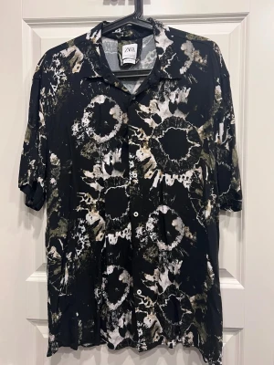 Svart mönstrad skjorta från Zara - Säljer en svart skjorta från Zara med avslappnad passform och unikt batikliknande mönster i vitt och olivgrönt. Skjortan har korta ärmar, klassisk krage och knäppning framtill. Perfekt för dig som vill sticka ut med en cool look.