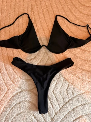 Svart bikini med v-formad topp - Säljer en stilren svart bikini med v-formad bygelbikinitopp och matchande tanga-bikinitrosa. Båda delarna har smala axelband och enkel design som ger en clean look. Perfekt för stranden eller poolen i sommar. Bikini överdelen är i L men är ganska liten för att vara en L och bikini underdelen är i M. Helt oanvända båda två. 