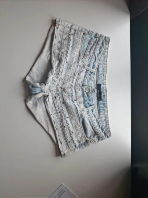 Ljusblå jeansshorts med spetsdetaljer - Unika ljusblå jeansshorts med flera spetsband och dekorativa knappar framtill. Klassisk femficksmodell och råa kanter nedtill för en trendig vibe. Perfekta för dig som vill sticka ut med en söt och cool stil. Materialet är denim med inslag av spets.