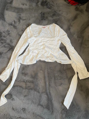 Vit topp med spets från Gina Tricot - Säljer en vit topp från Gina Tricot med långa ärmar och fina spetsdetaljer längs urringningen. Toppen har knytband i midjan och är tillverkad i mjuk bomull. Perfekt för dig som gillar en feminin och trendig stil.