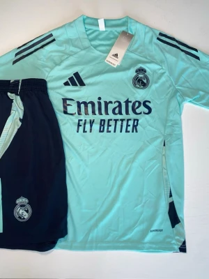 Real Madrid träningsset Adidas turkos - Säljer ett Real Madrid träningsset från Adidas i turkos och svart. Tröjan har klubbmärke, Adidas-logga och 'Emirates Fly Better' tryckt framtill. Matchande svarta shorts med klubbmärke. Tillverkad i lätt och ventilerande material, perfekt för fotbollsträning.