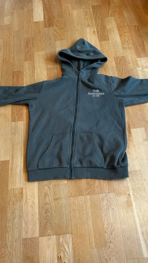 Mörkgrön hoodie Peak Performance - Mörkgrön  hoodie från Peak Performance med dragkedja framtill och två stora fickor. Klassisk huva och ribbade muddar vid ärmslut och nederkant. Diskret logga på bröstet. Perfekt för en avslappnad och sportig stil. Hör av er vid minsta fundering 😁