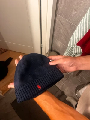 Mörkblå mössa från Polo Ralph Lauren - Mörkblå ribbstickad mössa från Polo Ralph Lauren med klassisk röd logga broderad framtill. Tillverkad i mjukt material som sitter skönt på huvudet och håller dig varm under kalla dagar. Enkel och stilren design som passar till det mesta.