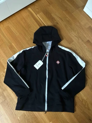 Casablanca zip-up Hoodie  - Snygg svart hoodie från Casablanca med vit dragkedja och vita ränder längs ärmarna. Har huva och detaljer i rött och vitt, inklusive logga på bröstet och dragkedjan. Tillverkad i mjukt bomullsmaterial som känns skönt mot huden.