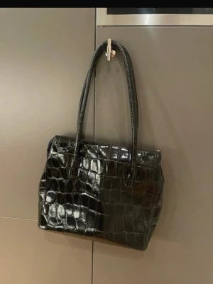 Vintage skinnväska i lackat krokodilmönster - En vintage skinnväska i lackat skinn med krokodilmönster och två fack inuti. Tidlös och jättefint skick!   Material: lackat skinn, tyg på insidan. Storlek: 30 x 35 cm (utan handtagen inräknat)