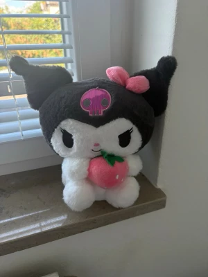 Kuromi mjukisdjur med jordgubbe - Söt Kuromi plushie i svart och vitt med rosa detaljer. Hon har en rosa rosett på ena örat, en rosa dödskalle på pannan och håller i en mjuk rosa jordgubbe. Perfekt för dig som gillar kawaii och samlar på Sanrio-prylar. 💗