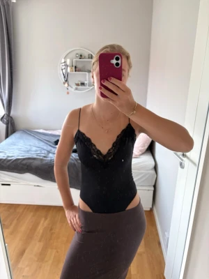 Svart spetsbody med tunna axelband - Snygg svart body med tunna axelband och djup V-ringning dekorerad med spetsdetaljer. Bodyn har en åtsittande passform och är tillverkad i ett stretchigt material som ger en smickrande siluett. Perfekt att styla med kjol eller jeans.