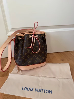 Helt ny Louis Vuitton monogram bucket bag i äkta läder - Helt ny handväska från Louis Vuitton i klassiskt brunt monogrammönster med beige och röda detaljer. Väskan har en bucket-form med dragsko och bred axelrem. Tillverkad i canvas med läderdetaljer. Perfekt för dig som gillar lyxiga accessoarer.