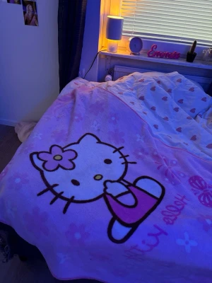 Hello Kitty rosa filt - Mysig rosa filt med stort Hello Kitty-motiv och blommor. Filtens material är mjukt och passar perfekt för att mysa i soffan eller ha som överkast. Motivet är i vitt, svart och rosa med texten 'Hello Kitty' och fjärilar.