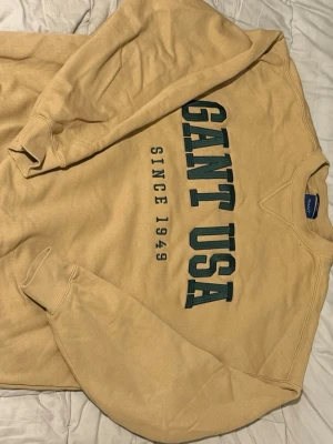 Beige sweatshirt från Gant - Säljer en beige sweatshirt från Gant med trycket 'GANT USA SINCE 1949' i grönt och svart på bröstet. Tröjan har rund halsringning, ribbade muddar och är tillverkad i mjukt bomullsmaterial. Perfekt för en avslappnad och stilren look.