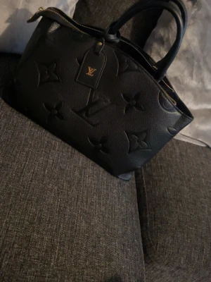 Svart Louis Vuitton handväska - Snygg svart handväska från Louis Vuitton i präglat läder med stora monogramdetaljer. Väskan har två handtag, guldfärgade dragkedjor och flera fack inuti för smart organisering. Perfekt accessoar för dig som gillar lyxig och tidlös design.