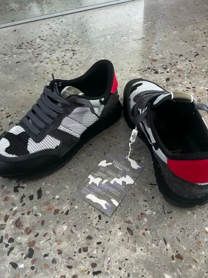 Valentino rockrunners helt ny - Snygga sneakers från Valentino med svart, grått och vitt camomönster samt en iögonfallande röd detalj på hälen. Skorna har svarta snören, mesh- och mockadetaljer samt en robust svart sula. Perfekta för dig som vill sticka ut med din stil.