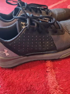 Svarta Under Armour sneakers - Snygga svarta sneakers från Under Armour med vita detaljer och prickigt mönster på sidan. Skorna har svart snörning, meshmaterial för bra ventilation och en låg profil som passar till många stilar. Perfekta för dig som gillar sportig och clean look.
