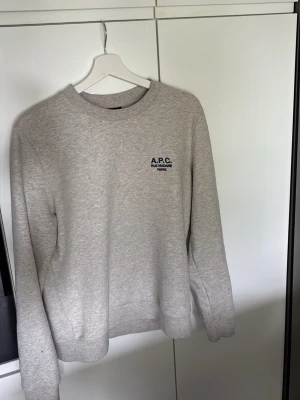 Ljusgrå sweatshirt från A.P.C. - Säljer en stilren ljusgrå sweatshirt från A.P.C. med diskret logga på bröstet. Tröjan har rund halsringning, långa ärmar och är tillverkad i mjukt bomullsmaterial. Perfekt för en clean och avslappnad look.