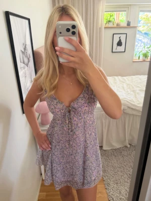 Ljuslila blommig kortklänning - Söt ljuslila kortklänning med blommigt mönster och volangdetaljer. Klänningen har smala axelband, v-ringning och en liten knytning framtill. Perfekt för varma dagar och har en luftig, flowy passform.
