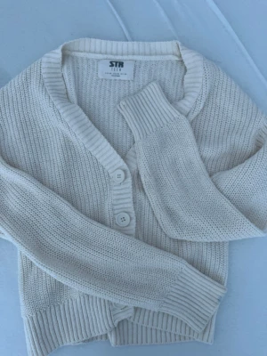 Krämvit stickad cardigan med knappar - Mysig krämvit stickad cardigan med två knappar framtill och breda ribbade muddar vid ärmslut och nederkant. Tröjan har en avslappnad passform och V-ringad hals. Perfekt att slänga över en t-shirt för en chill look.