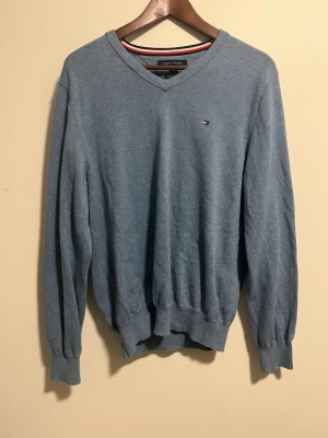 Blå stickad tröja Tommy Hilfiger - Snygg blå stickad tröja från Tommy Hilfiger med V-ringning och diskret logga på bröstet. Tröjan har långa ärmar och ribbade muddar vid ärmslut och nederkant. Perfekt för lager på lager och enkel att matcha.