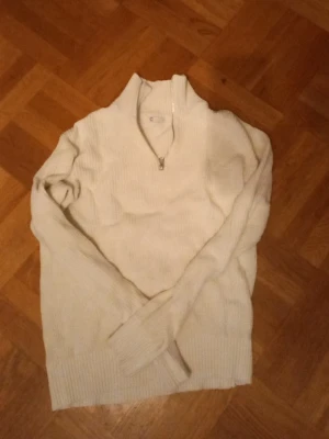 Vit stickad tröja med halv dragkedja - Klassisk vit stickad tröja med hög krage och halv dragkedja framtill. Tröjan har långa ärmar och ribbstickade muddar vid ärmslut och nederkant. Perfekt för lager på lager och enkel att matcha till olika outfits.