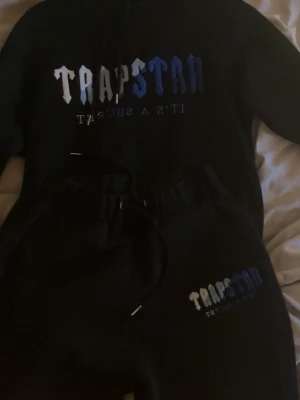 Trapstar svart loungewear set - Säljer ett svart loungewear set från Trapstar med hoodie och byxor. Setet har stor logga i vitt och blått på både tröjan och byxan samt texten 'It's a secret'. Hoodien har dragsko och byxorna har resår i midjan.
